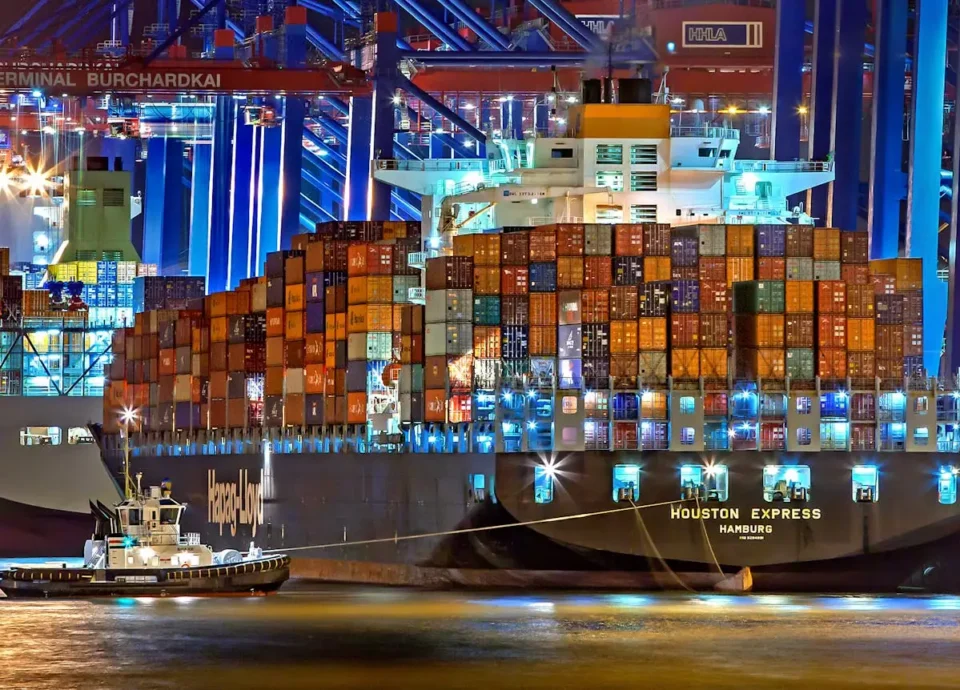 A brightly lit cargo ship at Hamburg harbor with stacked containers and a tugboat.